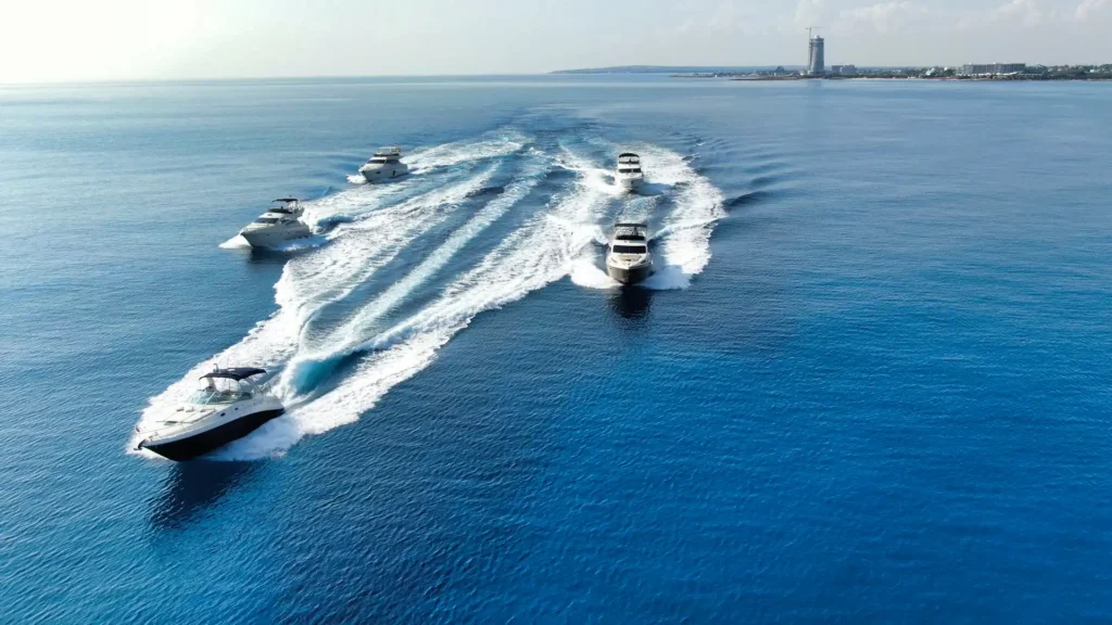 A fleet of yachts cruises in formation over the serene blue waters near the Ayia Napa coastline, creating symmetrical wakes.