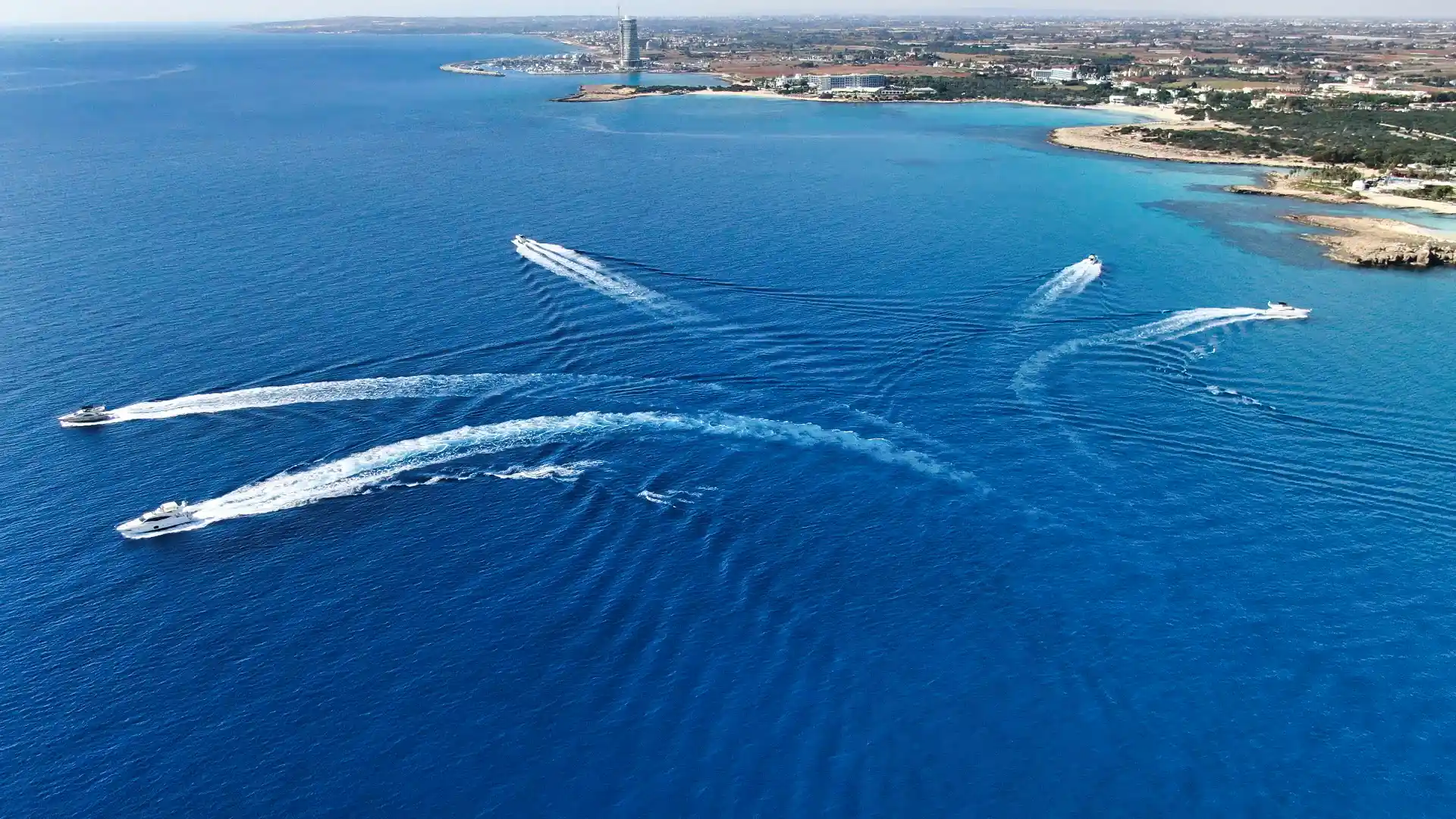 Yachts speeding through the sea near Ayia Napa, creating dynamic patterns on the water's surface with the coastline in the distance.