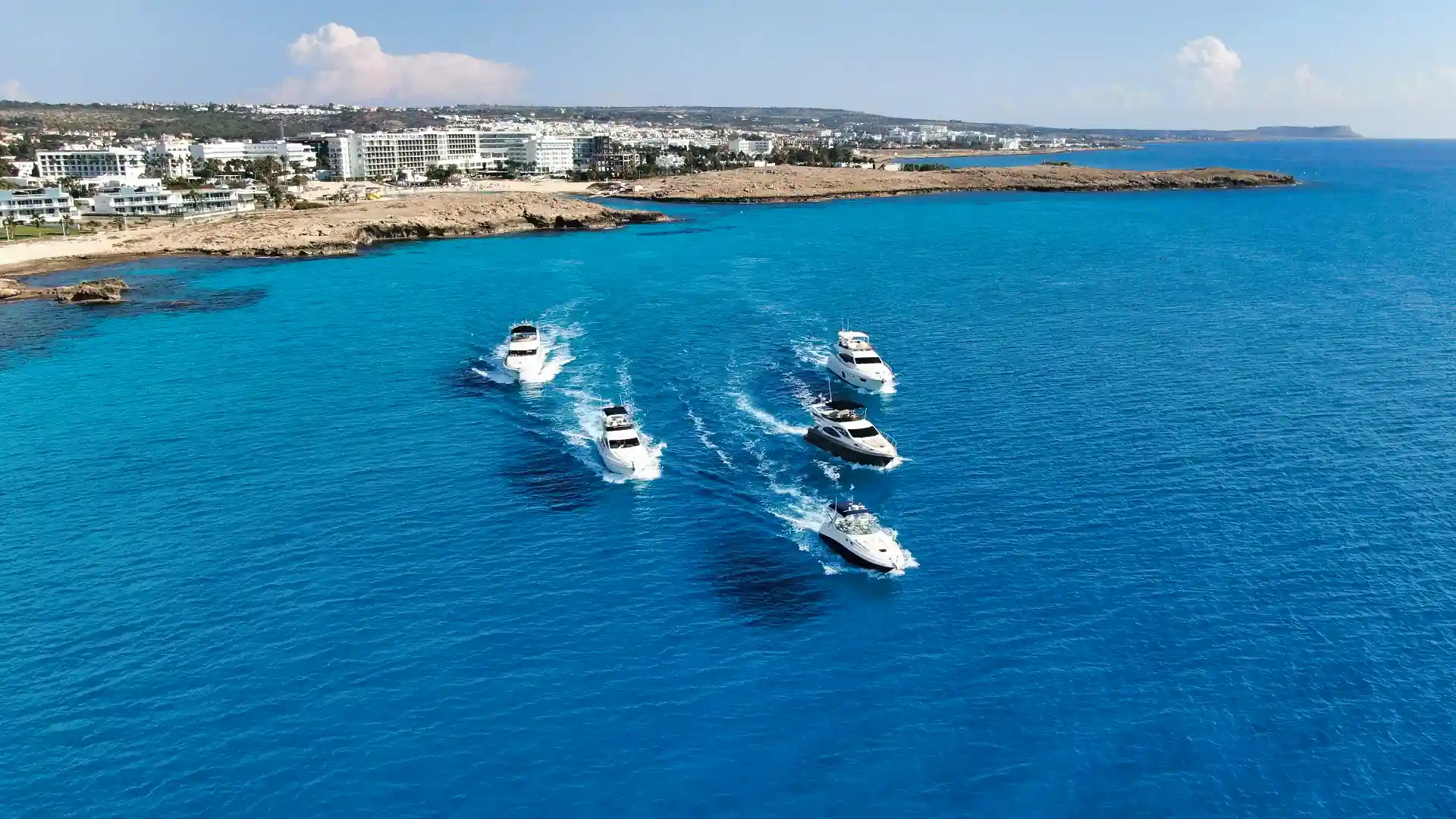 Aerial view of a group of boats cruising along the beautiful blue waters off the coast of Ayia Napa with resorts in the background.