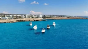 Leisure boats cruising on the turquoise waters near the rocky coastline of Ayia Napa, with resorts in the background under a clear sky.