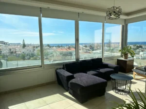 Spacious living room with large windows showcasing sea views, a dark sectional sofa, modern decor, and indoor plants in a sunlit Ayia Napa apartment.