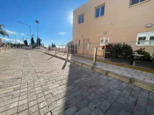 A sunlit paved pathway leading to a modern building, framed by greenery and clear skies in Ayia Napa.