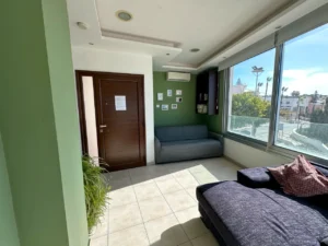 A contemporary living room with a green accent wall, gray sofa bed, and expansive windows with a city view in a bright Ayia Napa apartment.