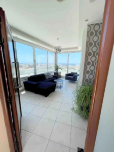 Entryway to a bright apartment with floor-to-ceiling windows overlooking Ayia Napa Bay and the city, with modern furnishings and greenery