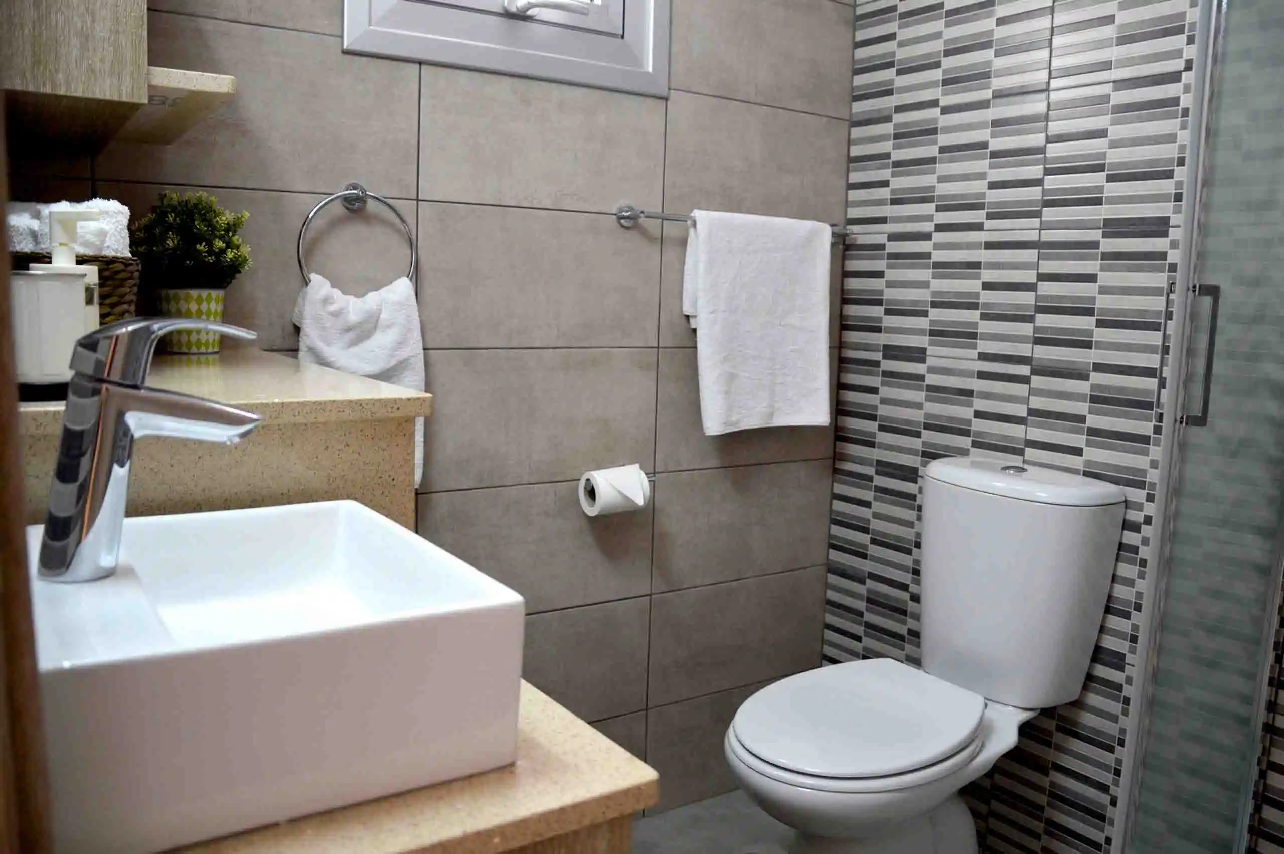 Modern bathroom with striped tile accent wall, rectangular basin, and wooden shelves, offering a refined and comfortable space in Ayia Napa.