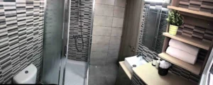 Wide view of a modern bathroom with walk-in shower, striped tiles, contemporary sink, and wooden shelves in an Ayia Napa apartment.