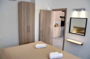 Bedroom view with a large wardrobe and a mirror reflecting the bed, with an open doorway leading to a well-equipped kitchen in a modern Ayia Napa apartment.