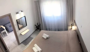 Bird's-eye view of a bedroom with natural light, sheer curtains, and a view into the living area with a black accent wall in a modern Ayia Napa apartment.