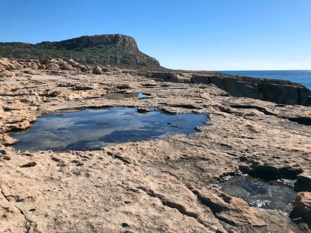 A serene view of the rocky landscape at Cape Greco near Ayia Napa, Cyprus, showcasing natural rock pools with clear water under a clear blue sky.