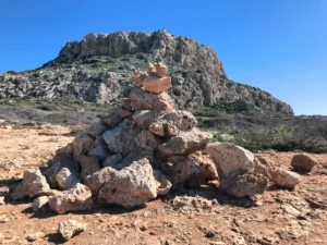 A cairn of balanced stones in the foreground with the rocky terrain of Cape Greco under a bright blue sky in the backdrop.