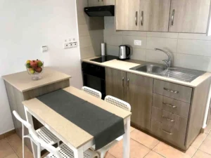 Modern kitchenette with wooden cabinets and a dining area, featuring a fruit bowl, in an Ayia Napa holiday home.