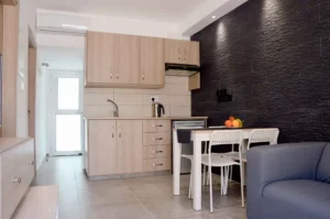 Modern kitchen with light wood cabinets and stainless steel appliances, adjacent to a dining area with white chairs and black textured wall in Ayia Napa apartment.