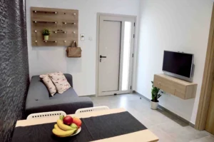 A cozy corner of a modern Ayia Napa apartment with a gray sofa, black textured wall, floating wooden shelf, TV, and a bowl of fresh fruit on a dining table.