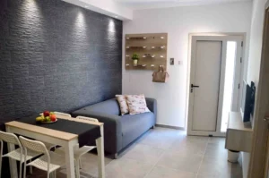 Modern Ayia Napa apartment living area with a dark textured accent wall, gray sofa, white dining chairs, and a wooden shelving unit.