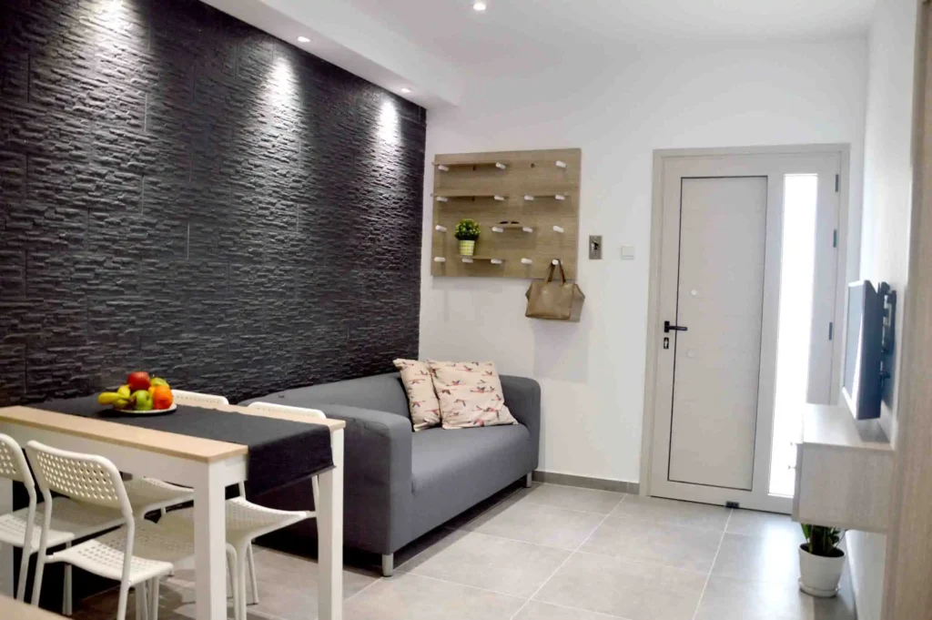 A chic and modern living room with a textured black accent wall, stylish gray sofa, white dining chairs, and a wooden shelf with decor, in an apartment in Ayia Napa, Cyprus.