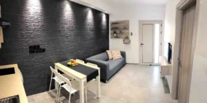 Panoramic view of a modern Ayia Napa apartment interior with black textured accent wall, gray sofa, white dining chairs, and a bowl of fresh fruit.