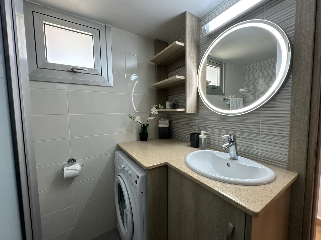 Bathroom with a sink, illuminated mirror, shelves, and a washing machine in the Charming Central Apartment with Sea View Balcony, Ayia Napa, Cyprus.