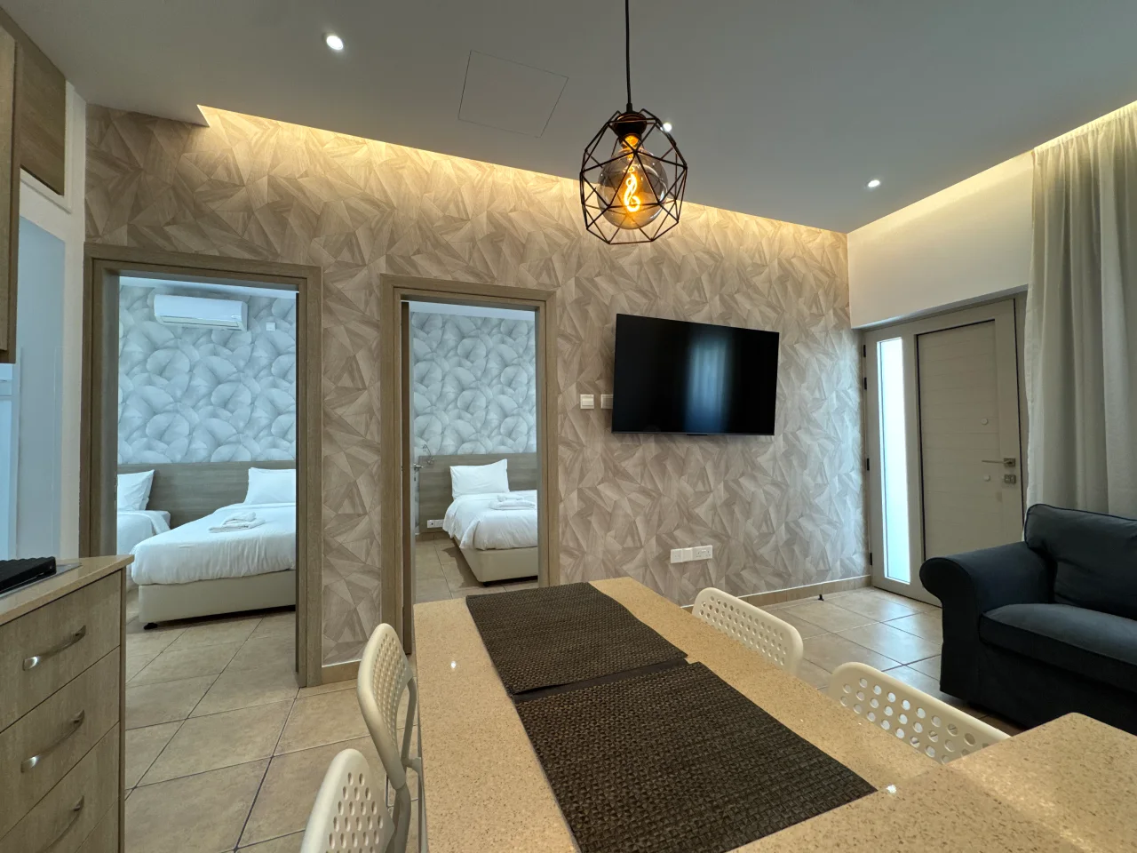 Bright living area with a sofa, TV, dining space, and doorways leading to the two bedrooms in our holiday apartment in Ayia Napa, Cyprus.